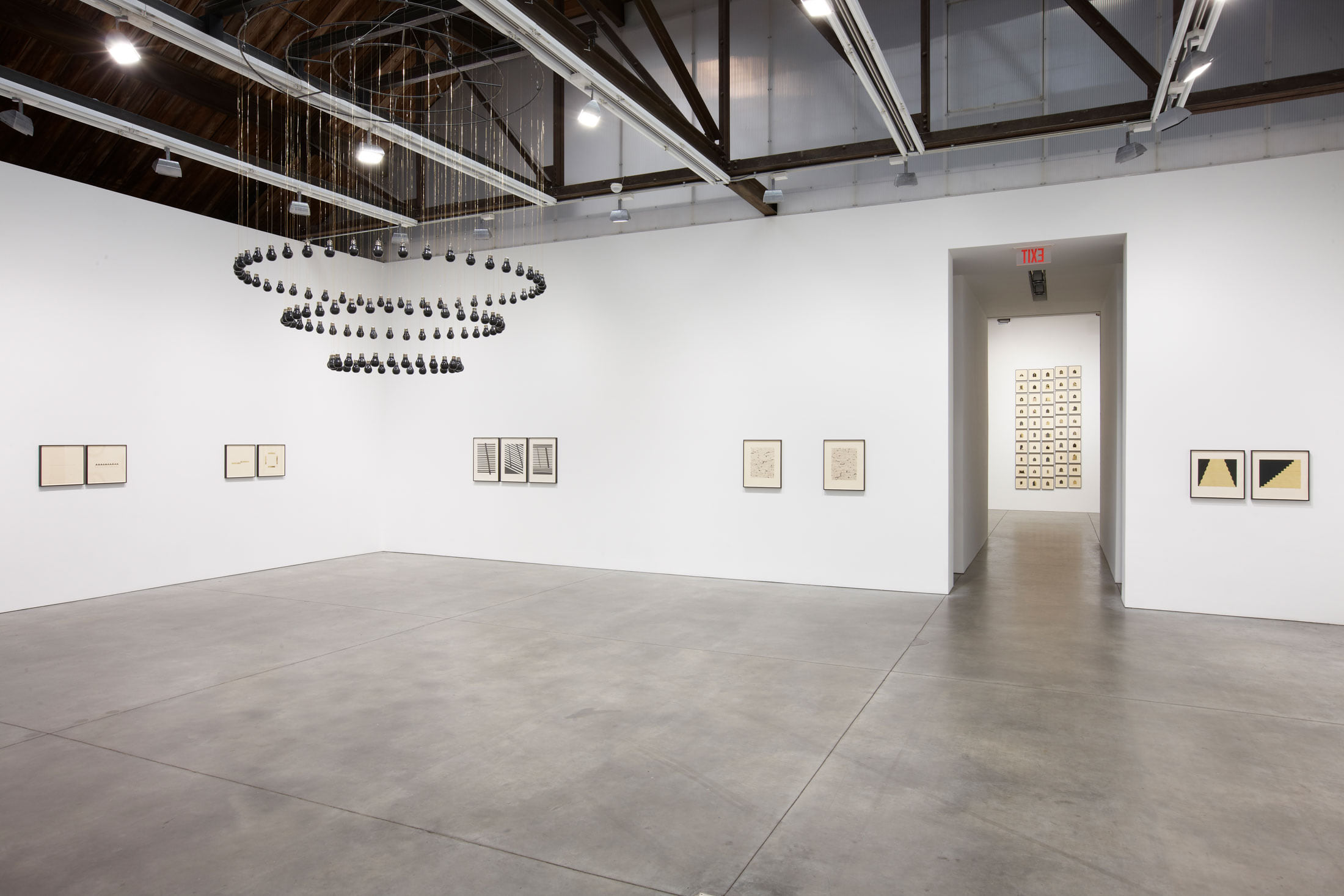 Installation view of Descending Darkness, with the suspended black-marble light bulbs of the title sculpture above works on paper